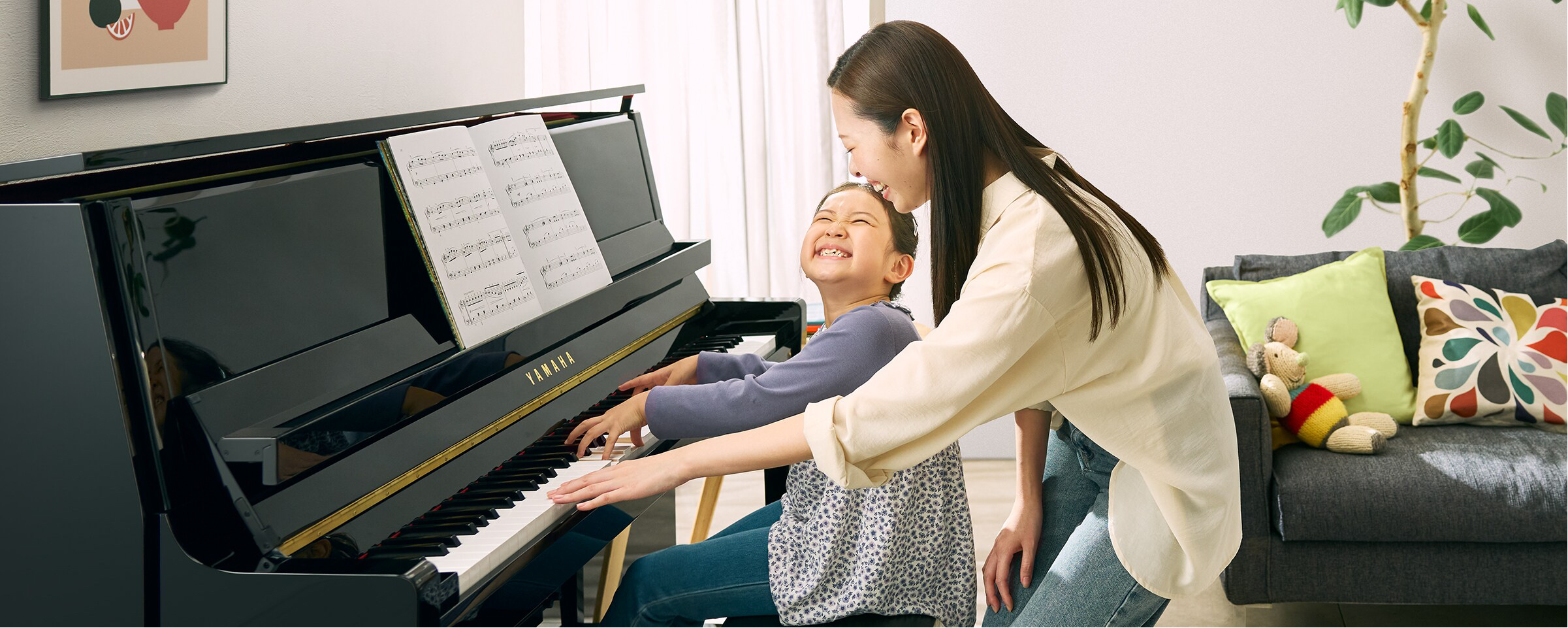 Mother helping daughter play a black Yamaha piano, both smiling as they press the keys together.