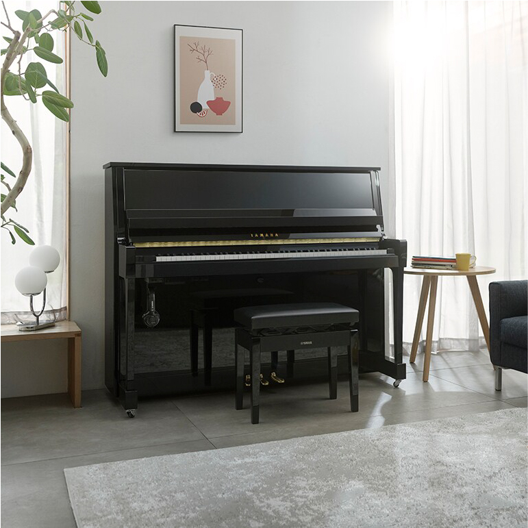 Black Yamaha upright piano with bench in a bright living room, plants and minimalist art on the wall nearby.