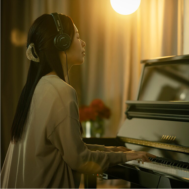 Young woman wearing headphones plays a grand piano in a warmly lit room, focused on the keys.