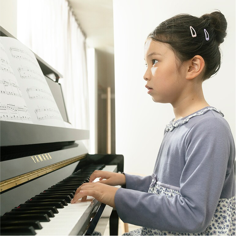 Young girl sits at a Yamaha piano, hands on the keys, reading sheet music as she practices.