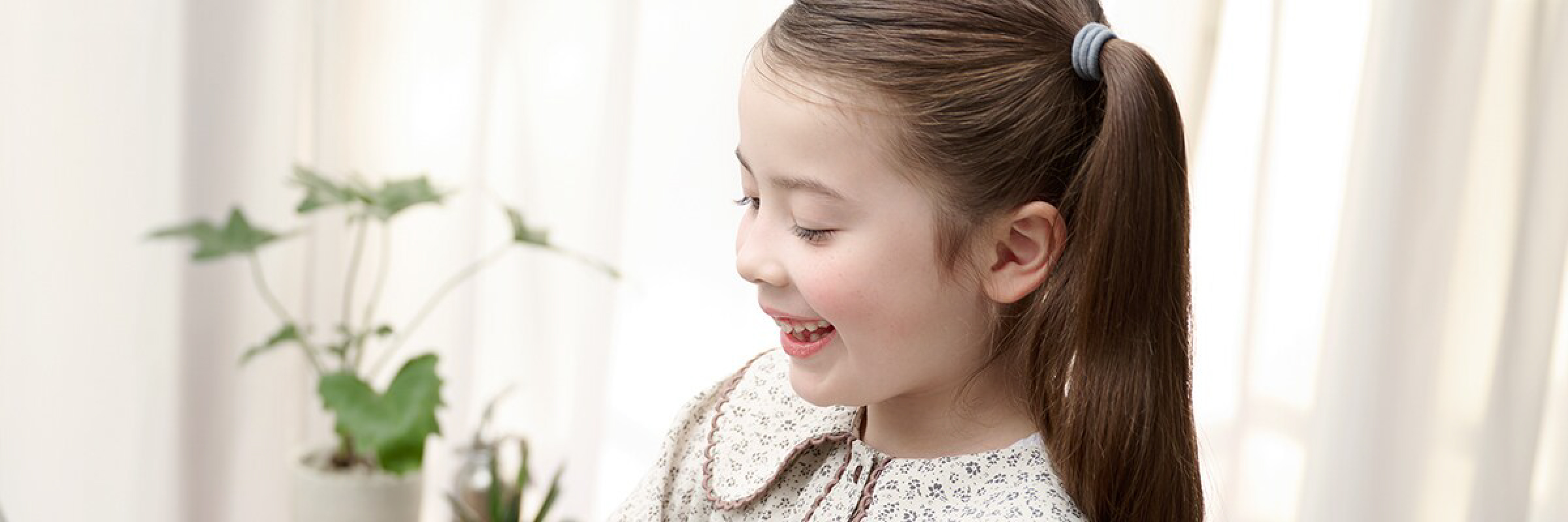 Side profile of a smiling young girl with long brown hair tied in a ponytail, indoors near a plant.