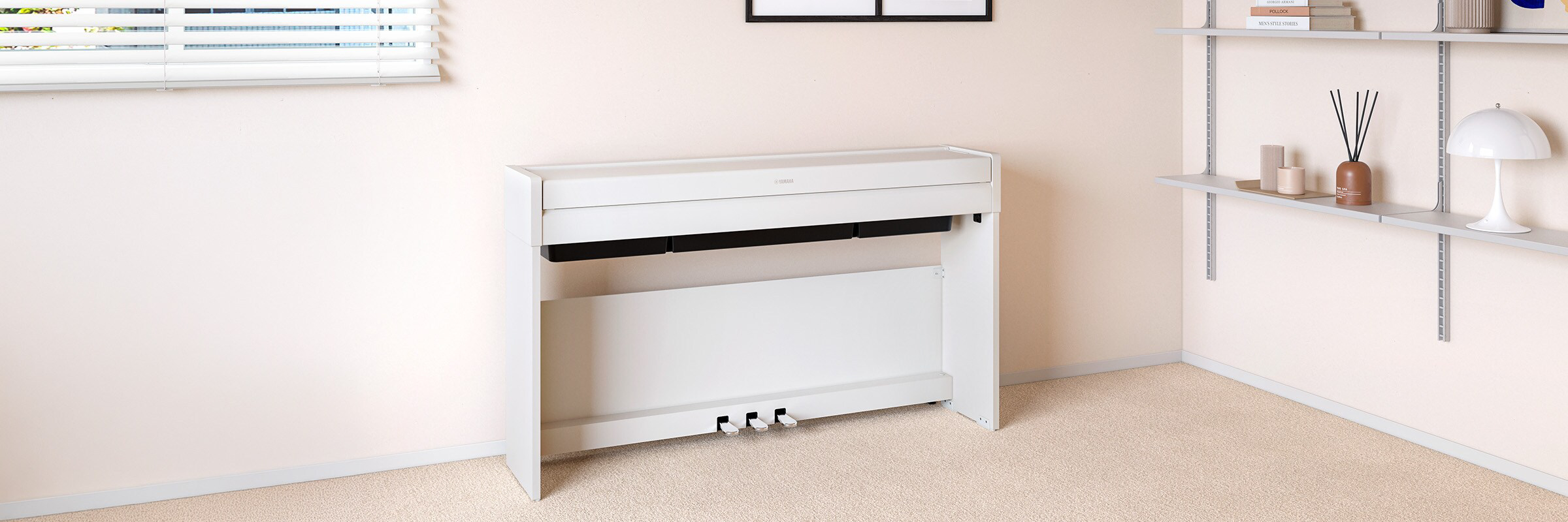 White Yamaha digital piano against a pale wall in a tidy room with blinds and shelving on the right, pedals visible at the base.