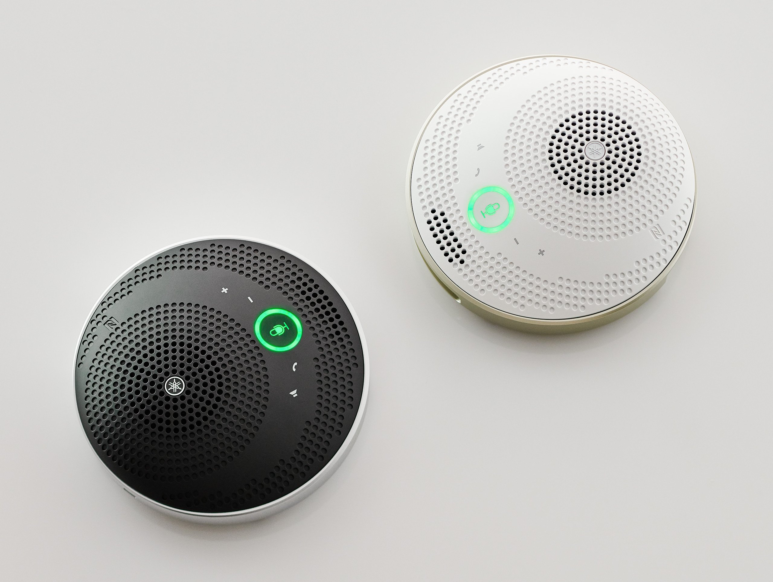 yvc-200_z_0003_e3fa711be7e340476861eb8497b9c58f - mustore two circular smart speakers with perforated tops, one black and one white, featuring green led rings indicating active audio. - mustore