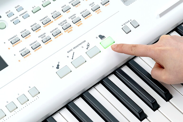 Close-up of a finger pressing a green button on a white electronic keyboard with many labeled controls and piano keys nearby.