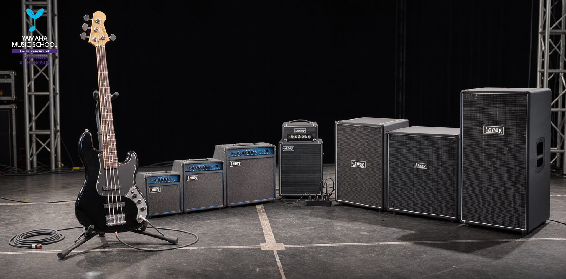 Stage with a black bass guitar on a stand next to a row of guitar amplifiers and speaker cabinets; coiled cable on the floor and stage lighting rig in the background.