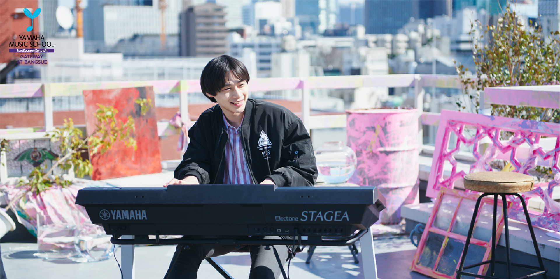 Young man playing a Yamaha Electone STAGEA keyboard on a rooftop with a city skyline in the background.