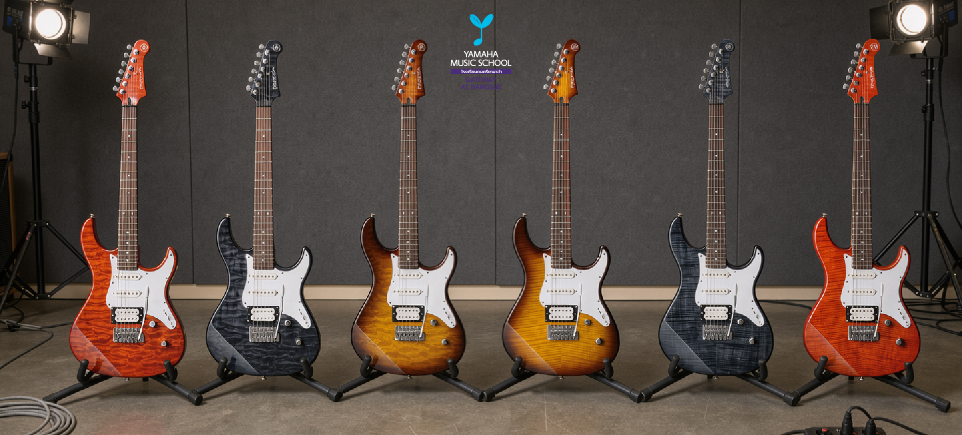 Six electric guitars on stands in a recording/rehearsal studio, with a Yamaha Music School logo centered above and cables on the floor ( finishes: red, black, sunburst).