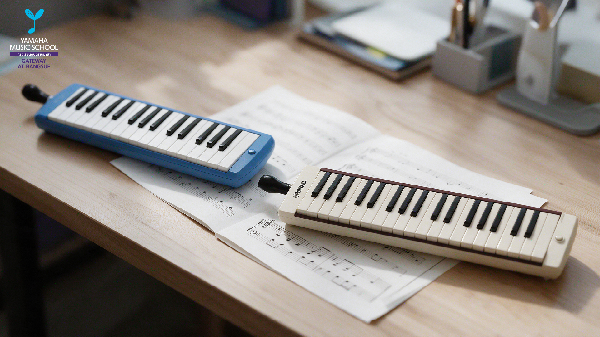 Blue Yamaha melodica and a beige melodica lie on a desk with scattered sheet music and a logo in the top left corner